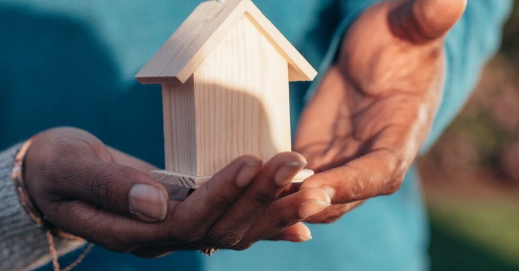 Close-up of hands holding a small wooden house model outdoors, symbolizing real estate investment.